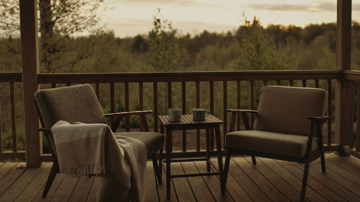 Two armchairs side by side on a wooden porch at golden hour, a folded blanket and two mugs on the small table between them