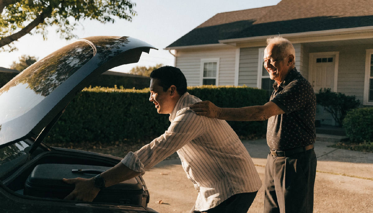 An adult son loading a suitcase into his car in a sunlit driveway while his elderly father stands beside him with a hand on his shoulder, both smiling