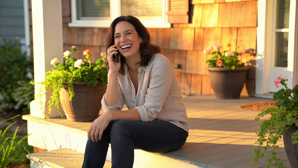 A woman in her thirties sitting on a sunlit front porch step, phone held to her ear, laughing mid-conversation
