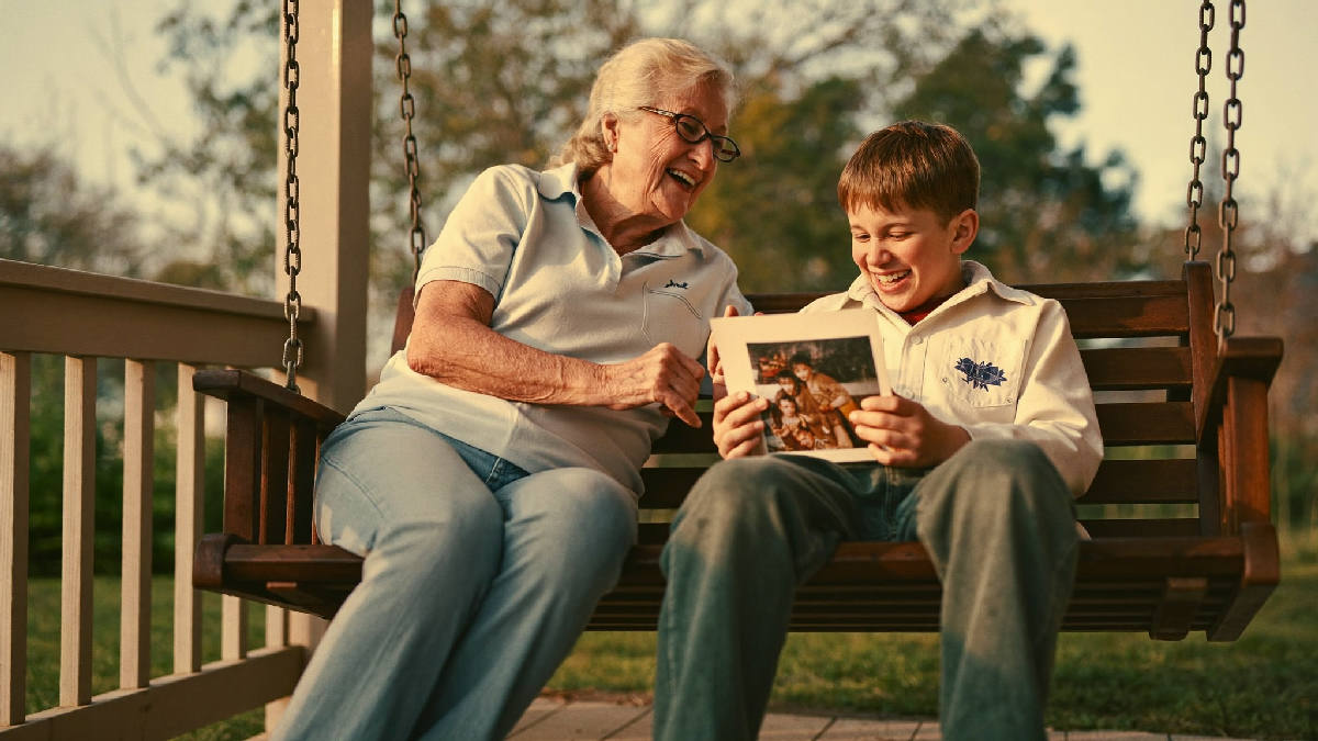 An elderly woman and her younger brother sitting together on a porch swing in warm late-afternoon light, laughing while looking at an old photograph