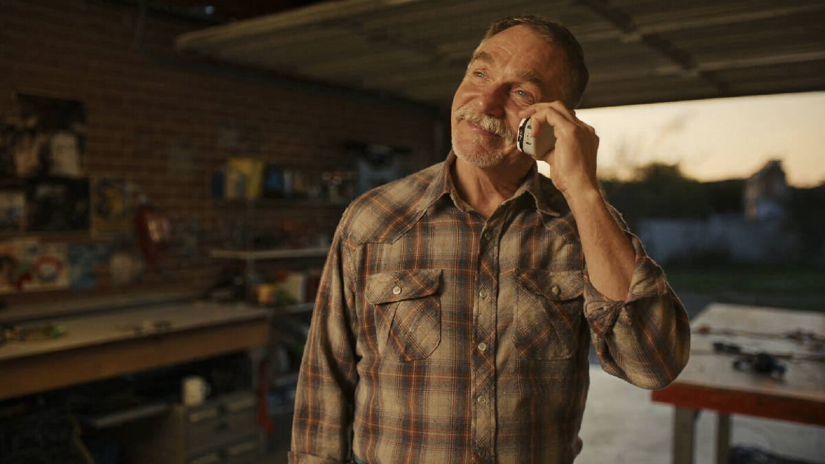 A man in his fifties in a plaid flannel shirt stands in his garage workshop at sunset, a phone to his ear, smiling softly as he listens