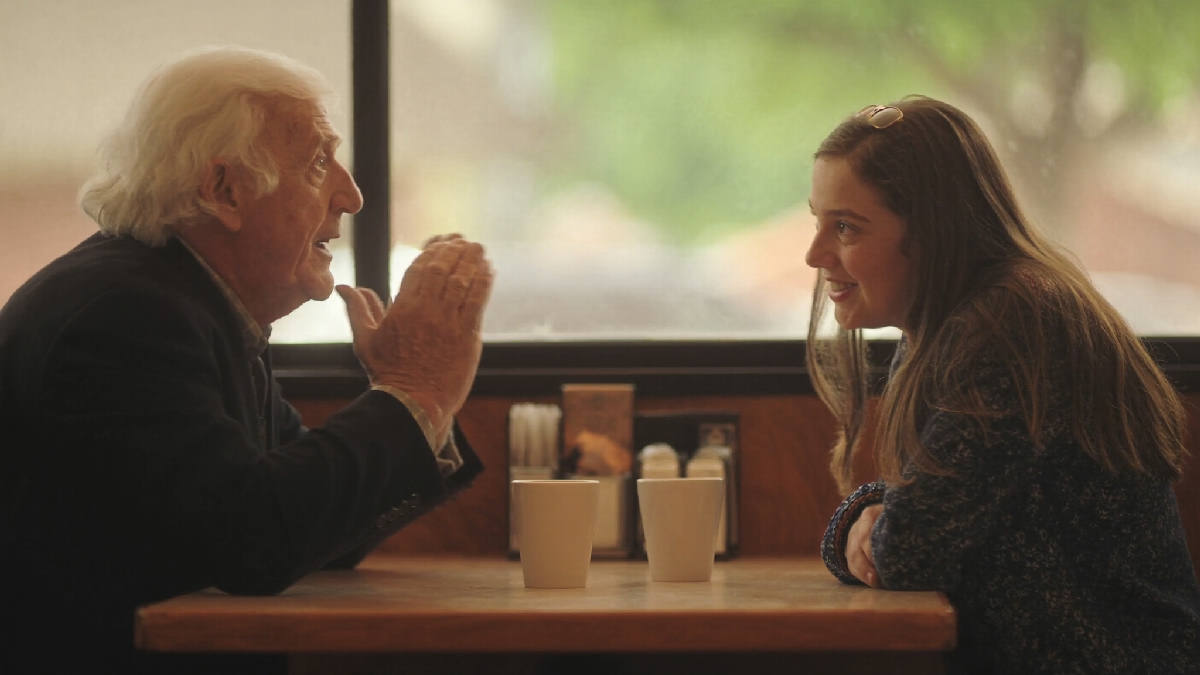 An elderly man with white hair tells a story with one hand raised across a diner booth table while his adult granddaughter leans forward, smiling, two coffee cups between them in warm afternoon light
