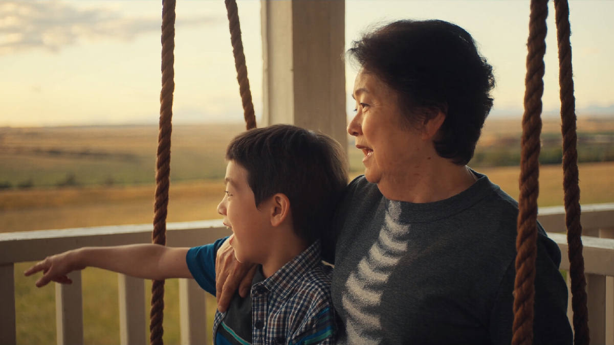 An older woman on a porch swing with her young grandson in late afternoon golden light, both looking out toward an open horizon, her arm around him