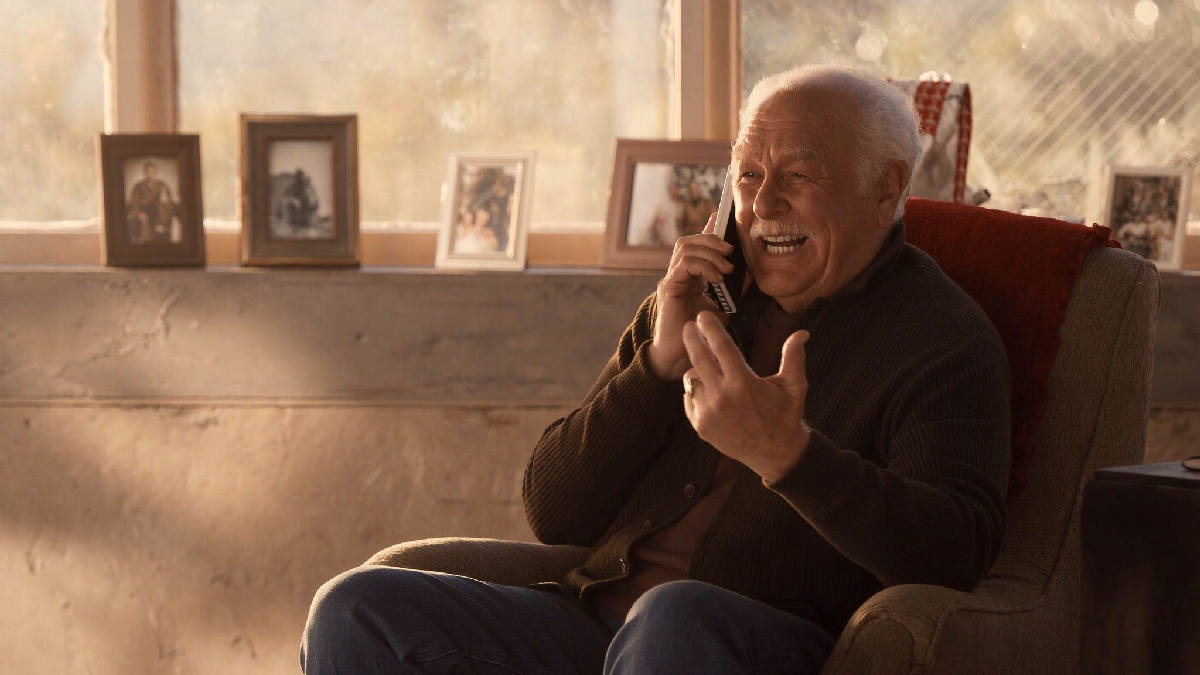 A man in his seventies sitting in a cozy armchair by a sunlit window, phone held to his ear, mid-laugh, framed family photographs on the wall behind him