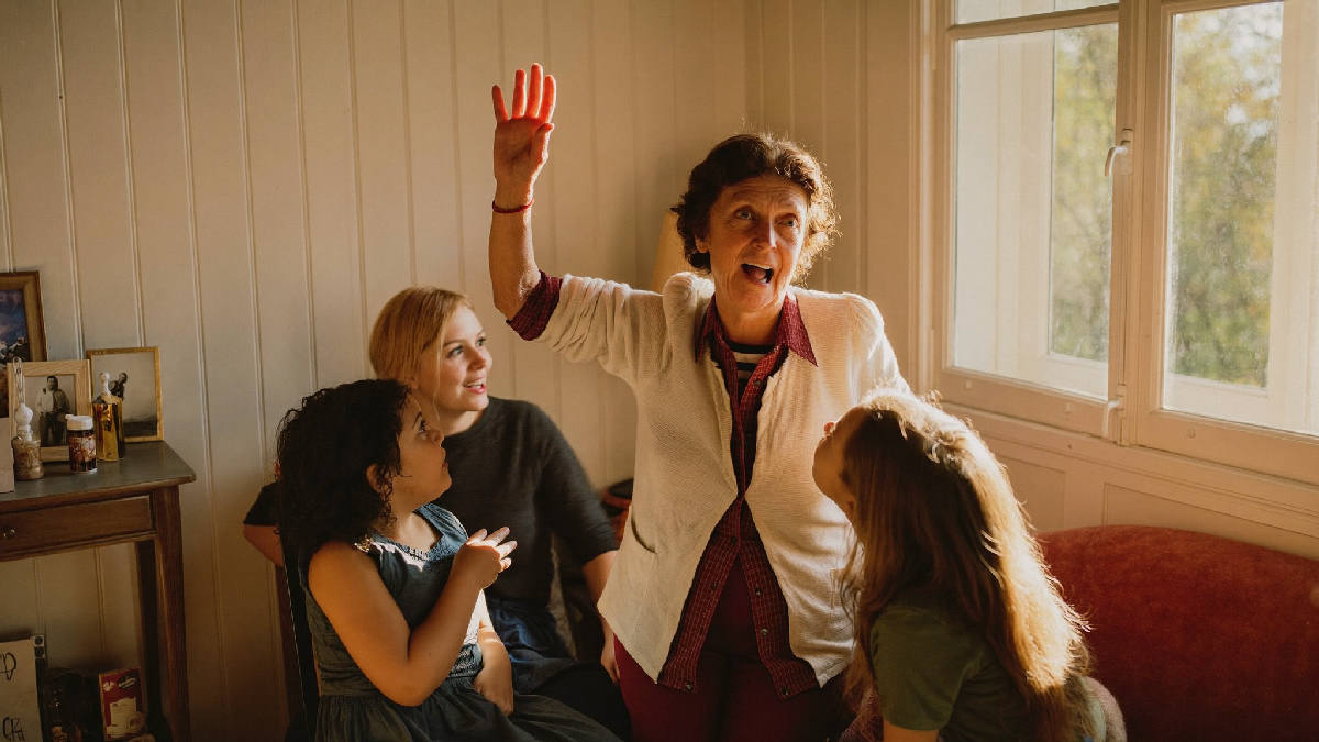 Three generations gathered in a sunlit living room, a grandmother mid-story with one hand raised, her adult daughter and grandchild leaning in and listening closely
