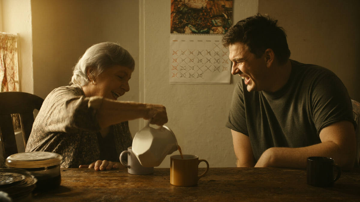 An elderly woman pours coffee and laughs with her adult son at a worn wooden kitchen table in golden morning light, a wall calendar of crossed-off days behind them