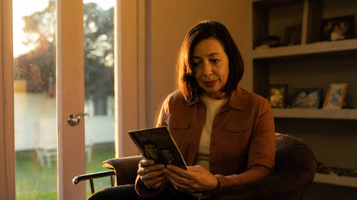 A woman in her mid-forties sitting alone by a sunlit window in the early evening, holding an old photograph, a soft content expression, family photo frames on a shelf behind her