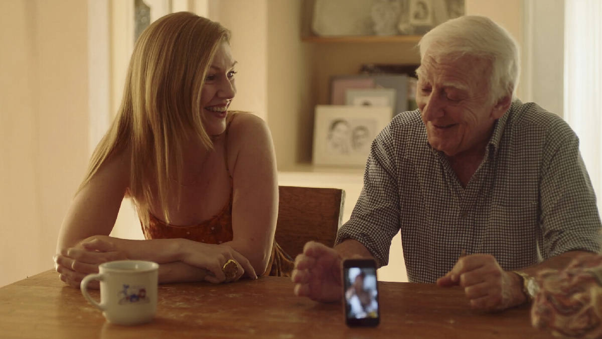 An adult daughter sitting close beside her elderly father at a sunlit kitchen table, a smartphone glowing softly on the table between them, both mid laugh in conversation